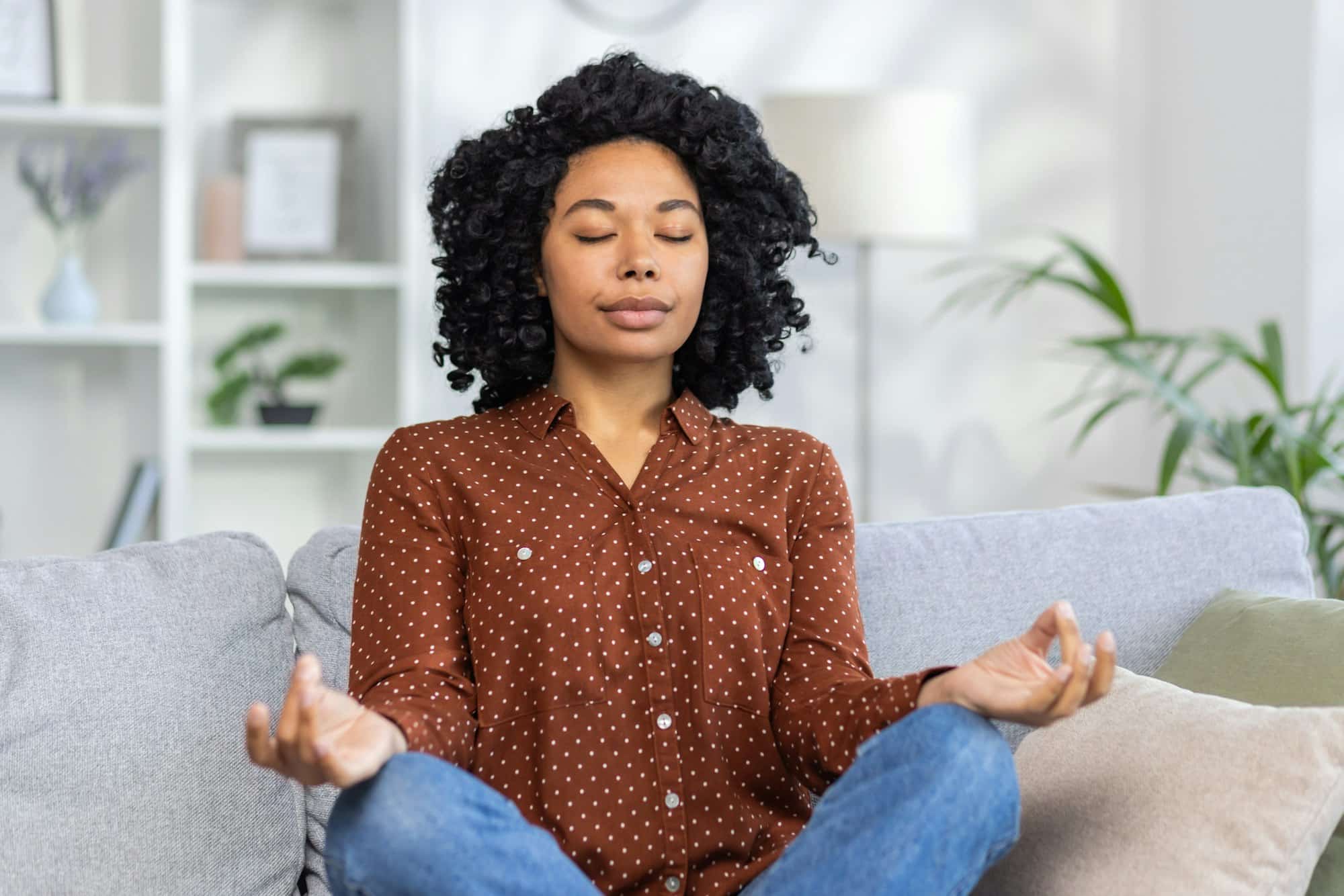 Serene african american woman meditating on couch for mindfulness and stress relief in comfortable